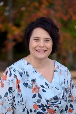  A woman with short, dark hair smiling at the camera in an outdoor setting with blurred autumn foliage in the background. She is wearing a light blue blouse with a floral pattern in orange, navy, and gray, along with a delicate necklace.