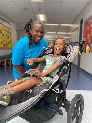 Female inclusion staff next to a male youth participant in a running stroller at a school based camp site.