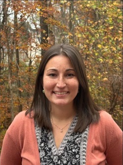 A woman smiling at the camera in an outdoor setting during autumn. She has shoulder-length brown hair, freckles, and is wearing a peach cardigan over a patterned black and white blouse. The background features trees with yellow and orange leaves.