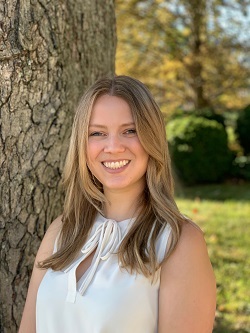 Caucasians, blonde female wearing a white sleeveless blouse standing in front of a tree.