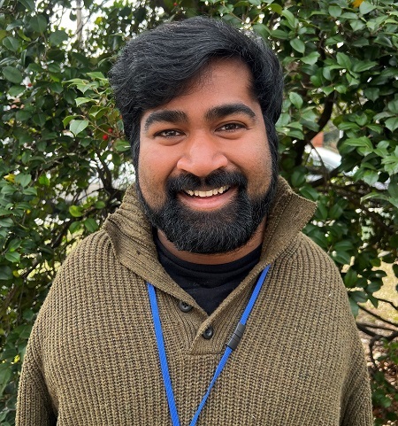 A man smiling with dark hair, a full bear, and brown skin, wearing a green sweater, standing in front of a tree